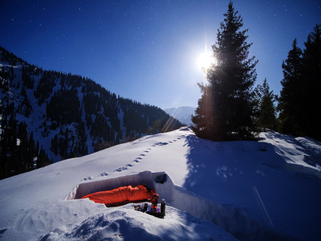 A male tourist sleeps in a sleeping bag in a snowy shelter on a moonlit winter night. Multi-day hikes in the winter mountains. Winter survival in the mountainsの写真素材