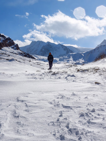 A male tourist is high in the snowy mountains. Bearded traveler in sunglasses with backpack and trekking poles. Cold, windy sunny day in the winter mountainsの写真素材