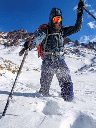 A male tourist walks knee-deep in deep snow high in the mountains. Bearded traveler with a backpack, trekking poles and wearing sunglasses.の写真素材