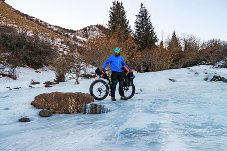 A bearded man travels in the mountains on a fat bike in winter. A man in a blue jacket and helmet stands with a bicycle on the ice of a frozen river. High mountain plateau Turgen-Asy, Kazakhstan.の写真素材