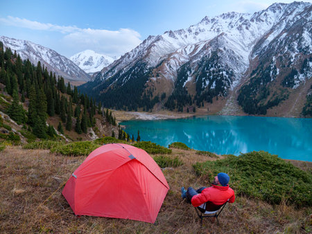 A male hiker sitting on a folding chair near a tent in a high mountainous area. He enjoys a magnificent panoramic view of snow-capped peaks and a turquoise mountain lakeの写真素材