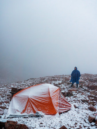 A hiker with a tent high in the mountains in a snowfall. Bearded man in blue raincoat and red tent during snowfallの写真素材