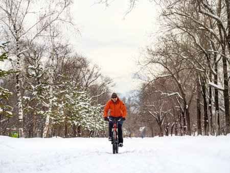 A man riding a bicycle in a winter park. A cyclist in an orange jacket rides on a snow-covered path. Ecological transportation in the cityの写真素材