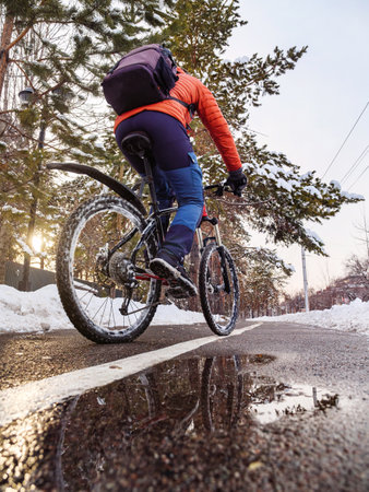 A man riding a bicycle on a bike path in winter in the city. Back view of a man in an orange jacket riding a bicycle. Ecological transportation in the cityの写真素材