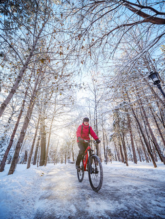 A cyclist is riding through a winter park in the early morning. The man is wearing a red jacket, helmet. Ecological transportation in the cityの写真素材