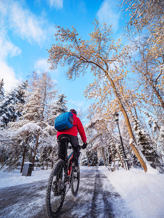 A guy riding a bicycle in a winter park in the early morning. The cyclist is wearing a red jacket, helmet and backpack. Rear viewの写真素材