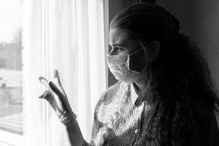 A young girl in a medical mask and gloves looks out the window while on self-isolation, quarantine at home. Home self-isolation in pandemic, flu epidemic, viral diseases, Covid-19.の写真素材