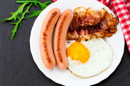 Breakfast with fried eggs, bacon, sausages and vegetable salad on dark stone background.の写真素材