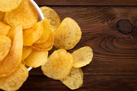 Crispy potato chips in bowl on wooden background, top view.