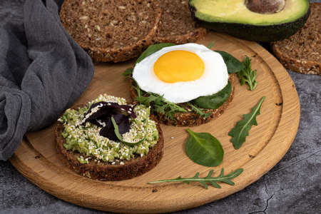 Avocado sandwich on dark rye bread with spinach and fried egg on a dark background.