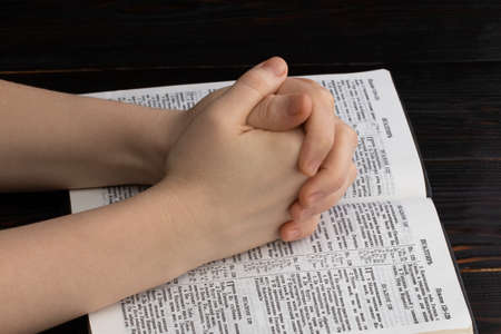 Hands praying with a bible in a dark over wooden table.