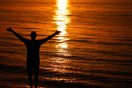 Silhouette of a man raising his hands or open hands as the sun rises by the sea.の写真素材