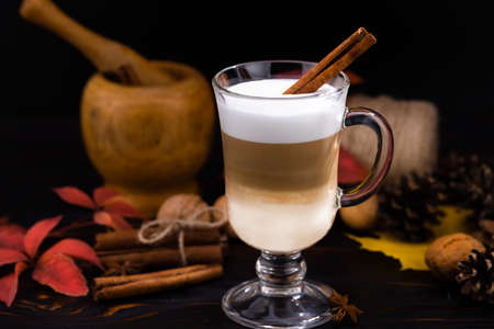 Glass cup of coffee latte and cinnamon sticks on a background of autumn leaves.
