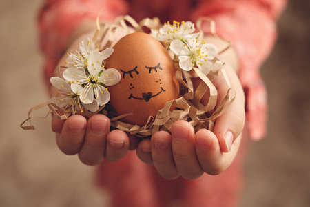 Easter egg with a face in the hands of a child decorated with blooming flowersの写真素材