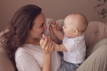 Portrait of a young mother and her one year old son at home. They play a funny game on the couch.