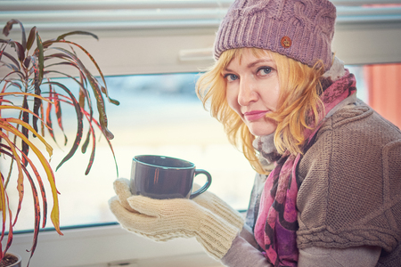 A woman in a wool cap and sweater with a Cup of tea looking out the window.の写真素材