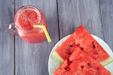 Chopped pieces of watermelon on a white plate and a glass mug with a watermelon smoothie on a gray tableの写真素材