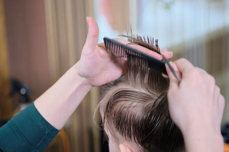 Barber shop. Hairdresser makes hairstyle to a  boy with hair scissors and black comb. Fashionable haircut for boys.の写真素材