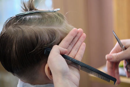 Barber shop. Hairdresser makes hairstyle to a  boy with hair scissors and black comb. Fashionable haircut for boys.の写真素材