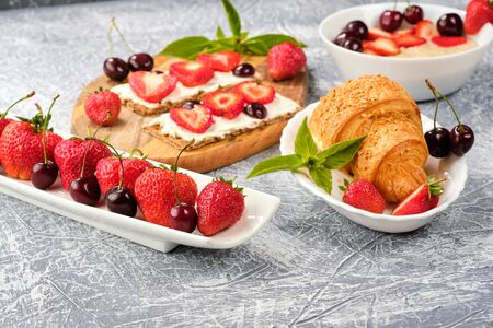 bowl of porridge with strawberries and cherries, croissants and crispbread with cheese and strawberries on a gray background. Healthy breakfastの写真素材