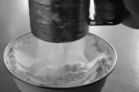Old stainless steel cup flour sifter in hand over a white enamel bowl. Monochrome image
