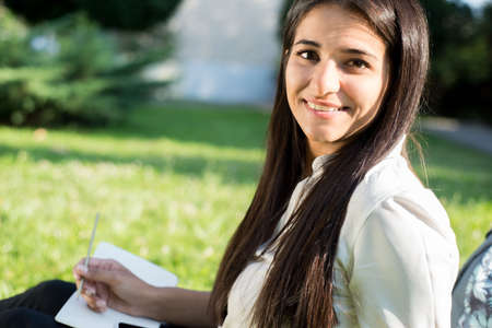 Portrait of a young Indian student, business woman. A woman with a backpack in a rucksack holds a tablet, sits on the green grass, against the sun. Summer dayの写真素材