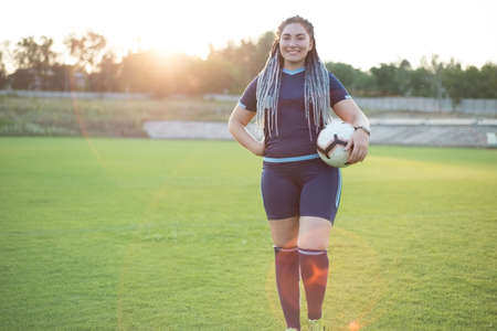 young girl with dreadlocks. In soccer uniform, holds the ball. Overweight woman, an alternative to sports. Losing weight. On the football field, green grassの写真素材