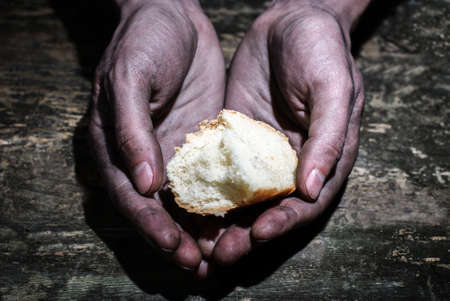 working dirty hands of a man holding a crust of bread on a dark old wooden table. Begging for a hand of hope. Give hope. The concept of alms and charity. Starving beggars ask for foodの写真素材