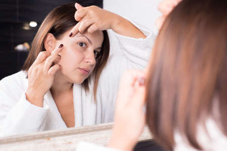 portrait of a beautiful girl, in the reflection of the mirror. Fingers remove acne on the face. Purulent folliculitis on the face. Boil, carbuncle. Facial skin care. A bad way to remove acne on the faceの写真素材