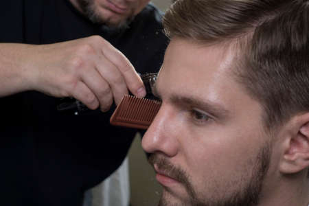 Hairdresser cuts hair with a trimmer for a young guy with a beard. Close-up of a master haircutting...