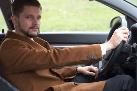 portrait of a young bearded guy in a suit driving a car. Business man in a coat and white shirt with...
