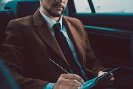 Closeup portrait of a young businessman with a beard. In stylish clothes, sits in the passenger seat, writing information in a notebook. In creative tinting. Business concept business style. A man rides in a taxi, working on the road. Doing business in a carの写真素材