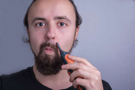 Close-up portrait of a young guy using a mustache trimmer. Beard and mustache care. A man trimmed...
