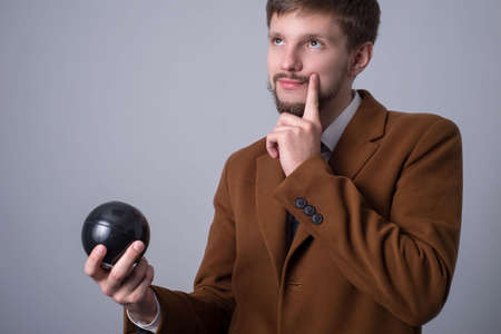 portrait of a bearded man in a business suit, holds a ball for making wishes, and asks a black billiard ball, holding his hand near his chin, dreams, looking up. On a gray background. Ball for making wishes from the film, track sixtyの写真素材
