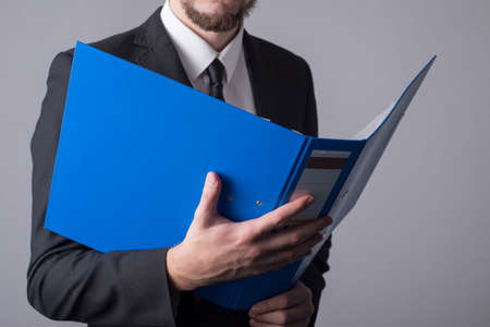 Portrait of a young bearded business man, with a blue folder for documents. On a gray background. Simple business concept, man businessman in suit studies business documents in business. using the paper folderの写真素材