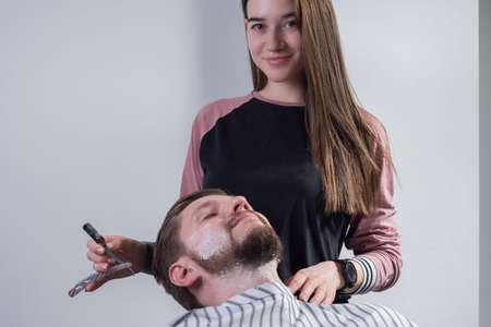 Close up process of haircut and beard care. A professional master prepares a clients beard for cutting and shaving a beard. The beauty of men. Barbers, beard and temples care salad. Portrait of a young girl, beard care professional, showing a dangerous razor. A male client sits nearby in an armchairの写真素材