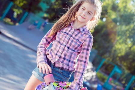 portrait of a little girl, in a plaid shirt, standing with a skateboard, on the playground. Teen...