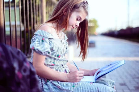 Portrait of a beautiful teenage girl with a backpack. Sitting on a bench writing in a notebook. Outdoorの写真素材