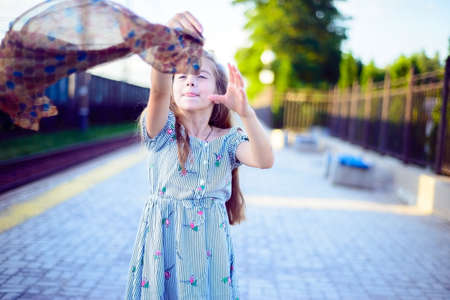 Portrait of a beautiful little girl in a dress playing with a fashionable scarf, tossing him in the air, and catches.の写真素材