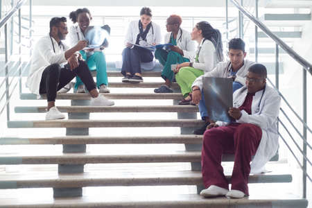 young doctors, mixed race, sitting, standing on the steps, in the corridor of the hospital. They consider x-rays, use gadgets to work in medicine. People in medical clothes.の写真素材