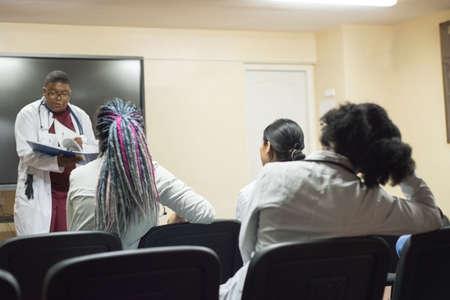 doctors, mixed race, in the classroom, at a medical conference. Young people, in white coats, with stethoscopes, are studying medicine. Classroom, anatomical museum. Students raise their hand to answer the question of the teacher, professorの写真素材
