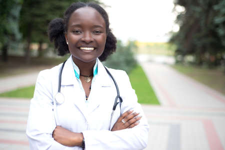 portrait of a young beautiful african girl, doctor. Woman in white coat with a stethoscope smiling. Outdoor inの写真素材