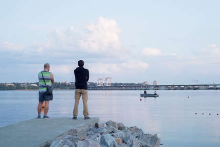 two people are standing on the pier, near the river. Watching the fishermen catch fish from a boat in the river.の写真素材