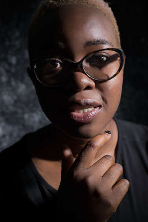 portrait of an african girl in glasses. On a dark background, artistic light. Woman holds hand at chinの写真素材
