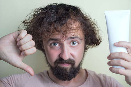 Portrait of a young guy with dirty hair and problematic scalp. Holding bad shampoo in hand, showing thumb down. Studio shooting. Ineffective shampoo, a hair care product for menの写真素材