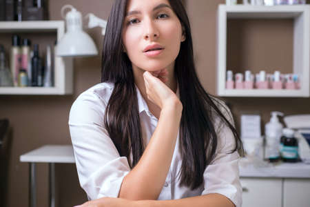 portrait of a young beautiful girl of twenty-five, in a white shirt, sitting at a table, taking elegant hand poses. Woman seller of cosmetics in a beauty salon. Manicurist. Young girl beautician in a beauty salon waiting for a client. Chewing gum beautician at workの写真素材