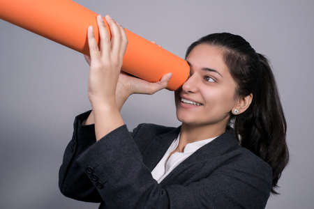 Portrait of a beautiful Indian young business woman in a business suit, holding a twisted orange tube in her hand, looking into it. Free place. Studio photography. On a gray background. Business concept Office style, business fashion. Woman at work, business woman in a business suit. A look into the future. Girl looks at her success.の写真素材