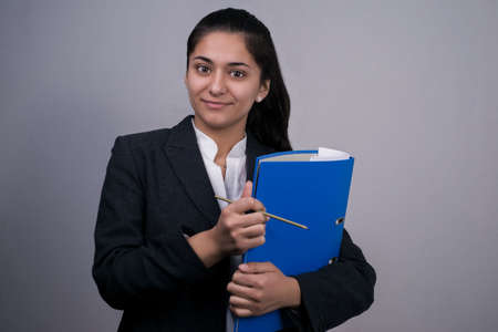 Portrait of a beautiful Indian young business woman in a business suit, holding a blue folder with documents in her hand, looking thoughtfully, Free space Studio photography. On a gray background. Business concept Office style, business fashion. Woman at work, business woman in a business suit.の写真素材