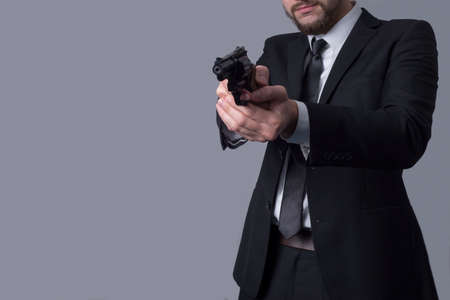 Portrait of a bearded man in a business suit holds an aiming revolver. On a gray background. Criminal type man, gangster, killer, businessman killerの写真素材