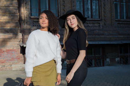 A pair of beautiful Mexican girls standing against the backdrop of an old university building. Two sisters. Young female students. Fashion shooting. Girl with curly hair, a white sweater and a leather jacket. Girlfriend in black clothes and a hatの写真素材